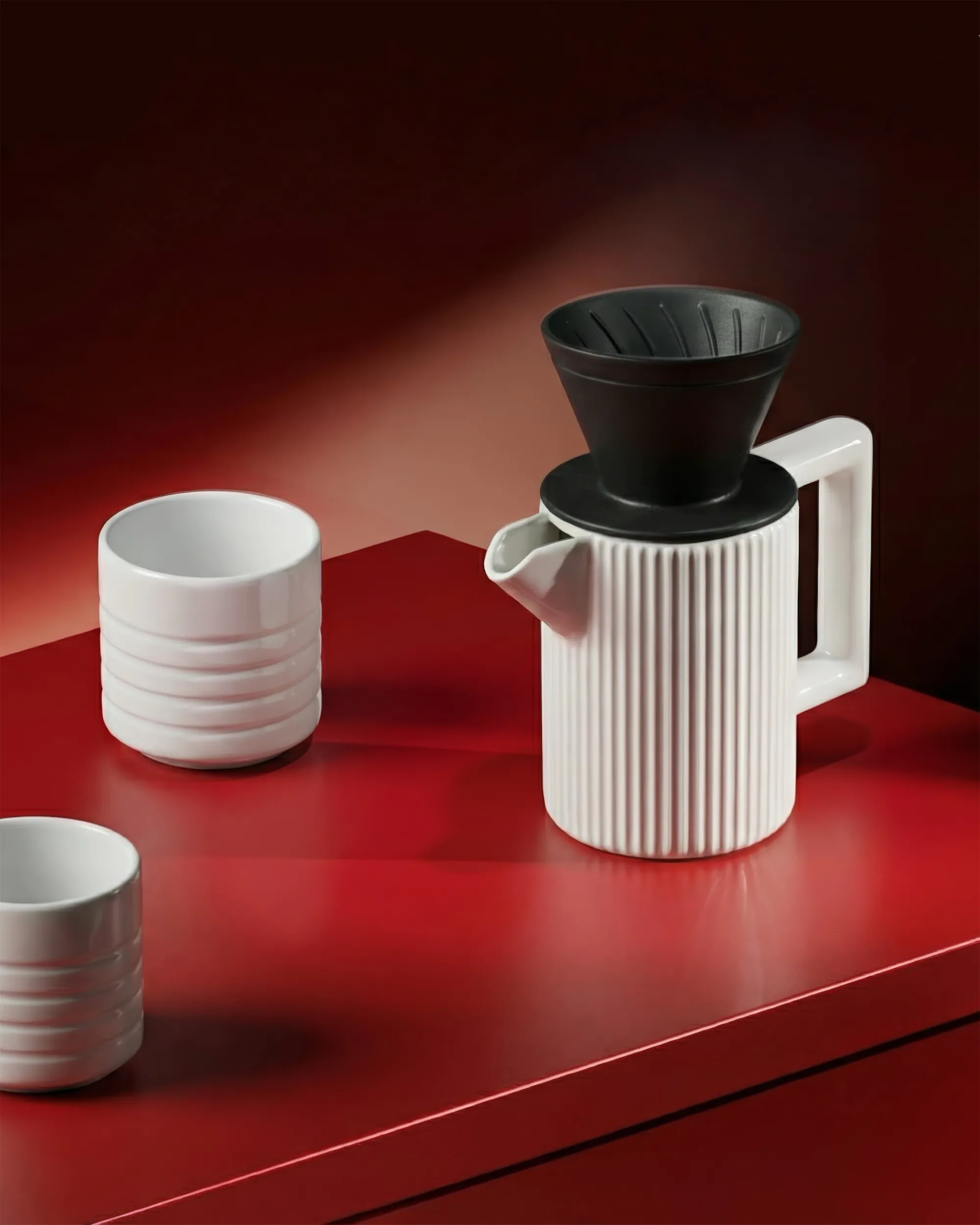 A white ceramic coffee maker and cup on a red table in a dimly lit room.