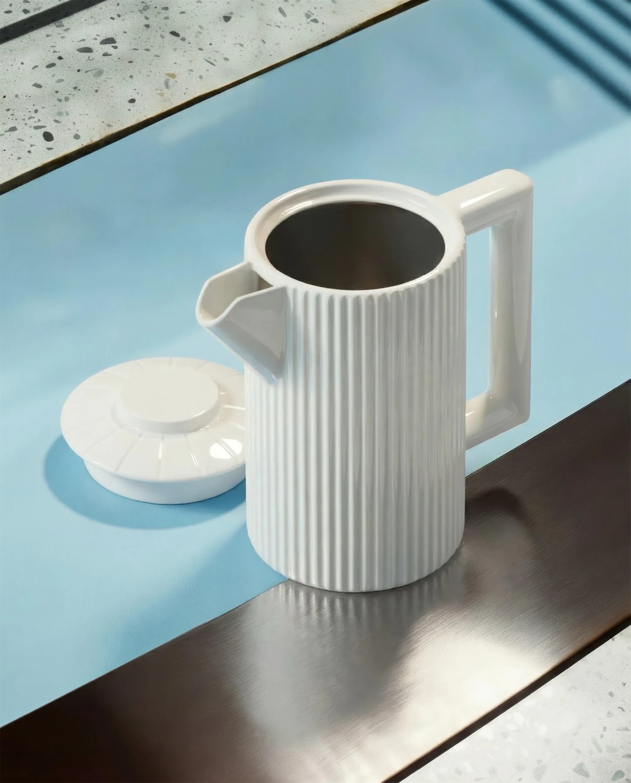 Белый керамический кофейник с ручкой и крышкой на светло-синем столе.