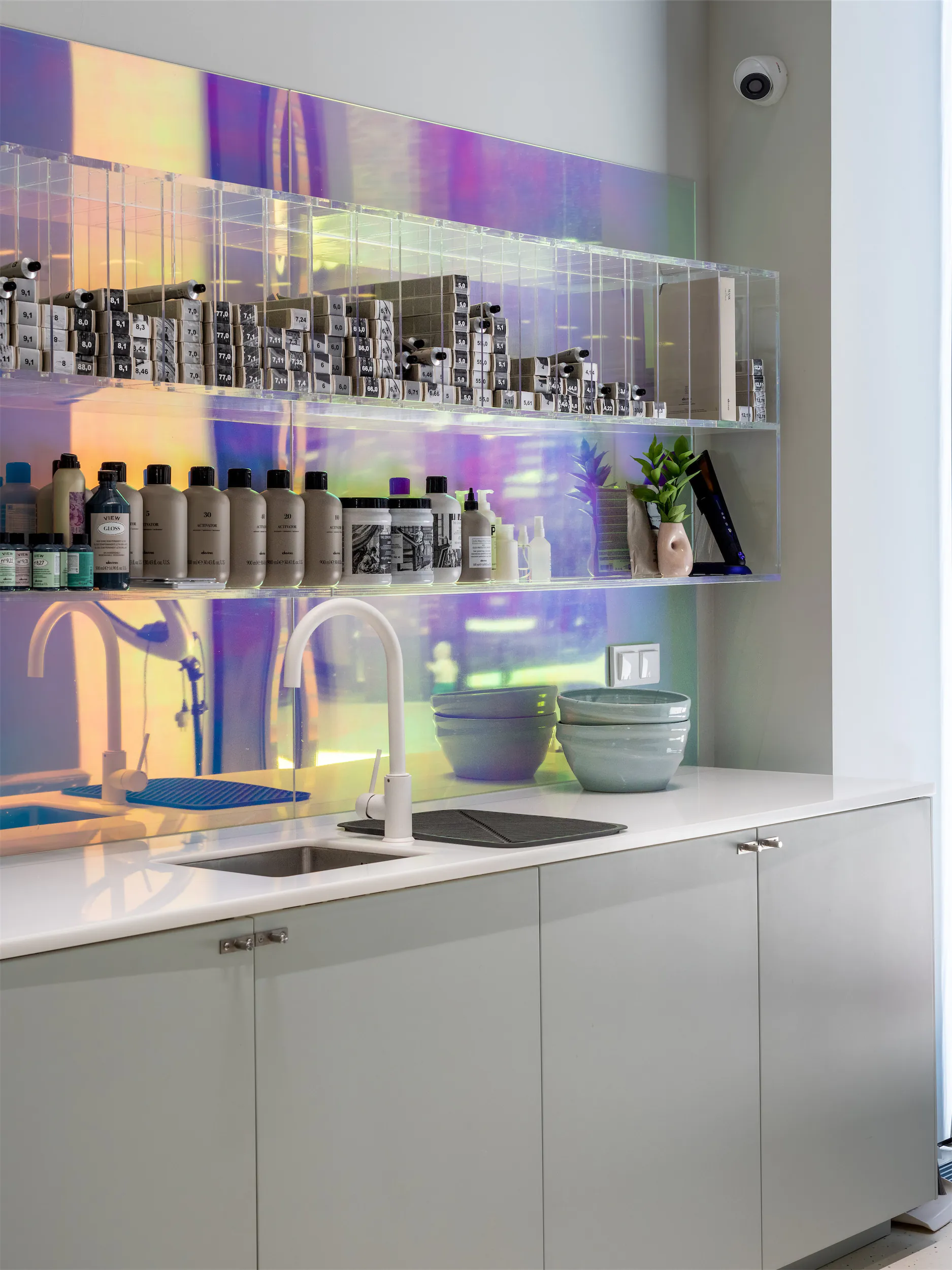 Белая раковина с краном под навесной полкой. На столешнице стоят флаконы.