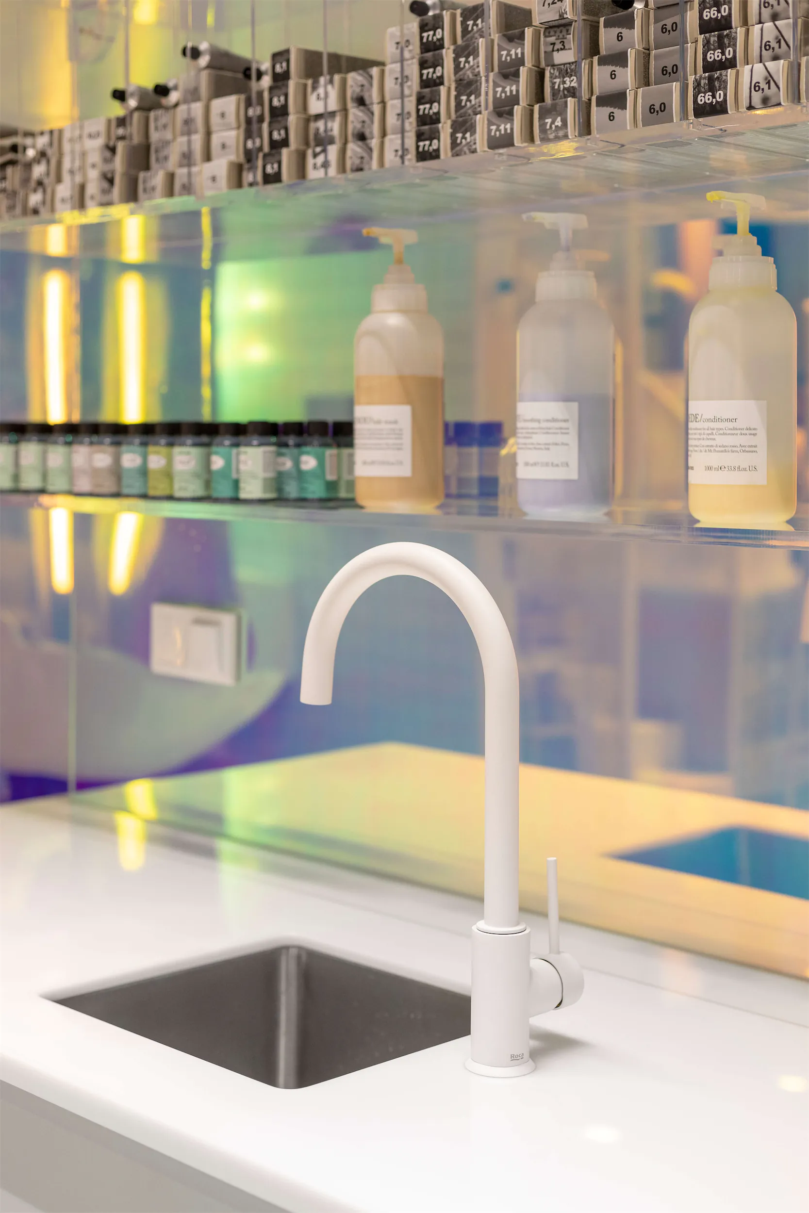 Белая раковина с изогнутым краном на белой столешнице рядом с полкой с флаконами и контейнерами.