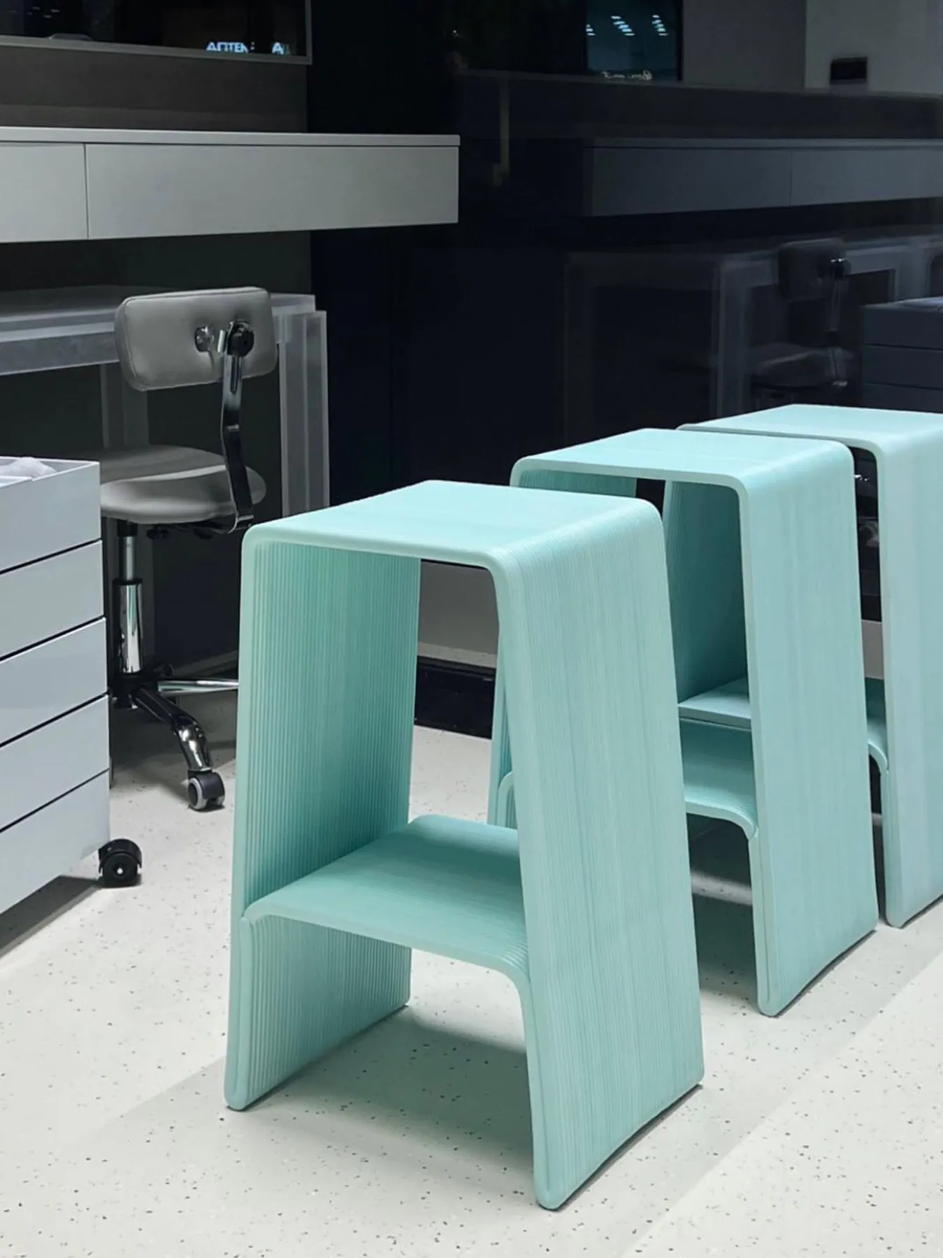 Два бледно-голубых пластиковых табурета на белом полу перед серым креслом и тумбой с ящиками.