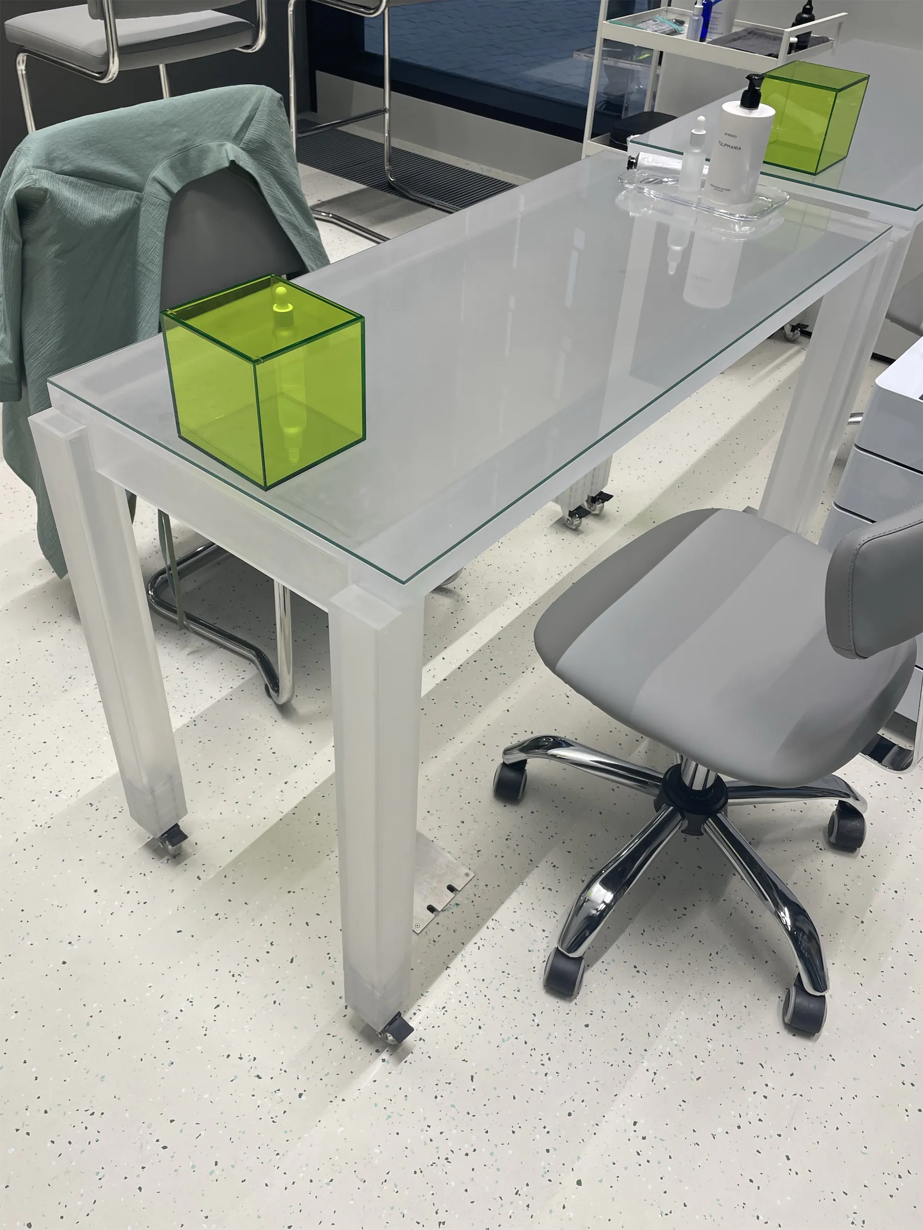 Белый стол со стеклянной столешницей и серое офисное кресло на полу с крапинками.