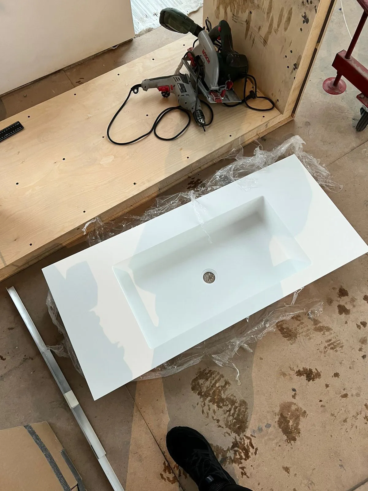 Белая прямоугольная раковина на полу среди частично собранного деревянного каркаса и электроинструмента.