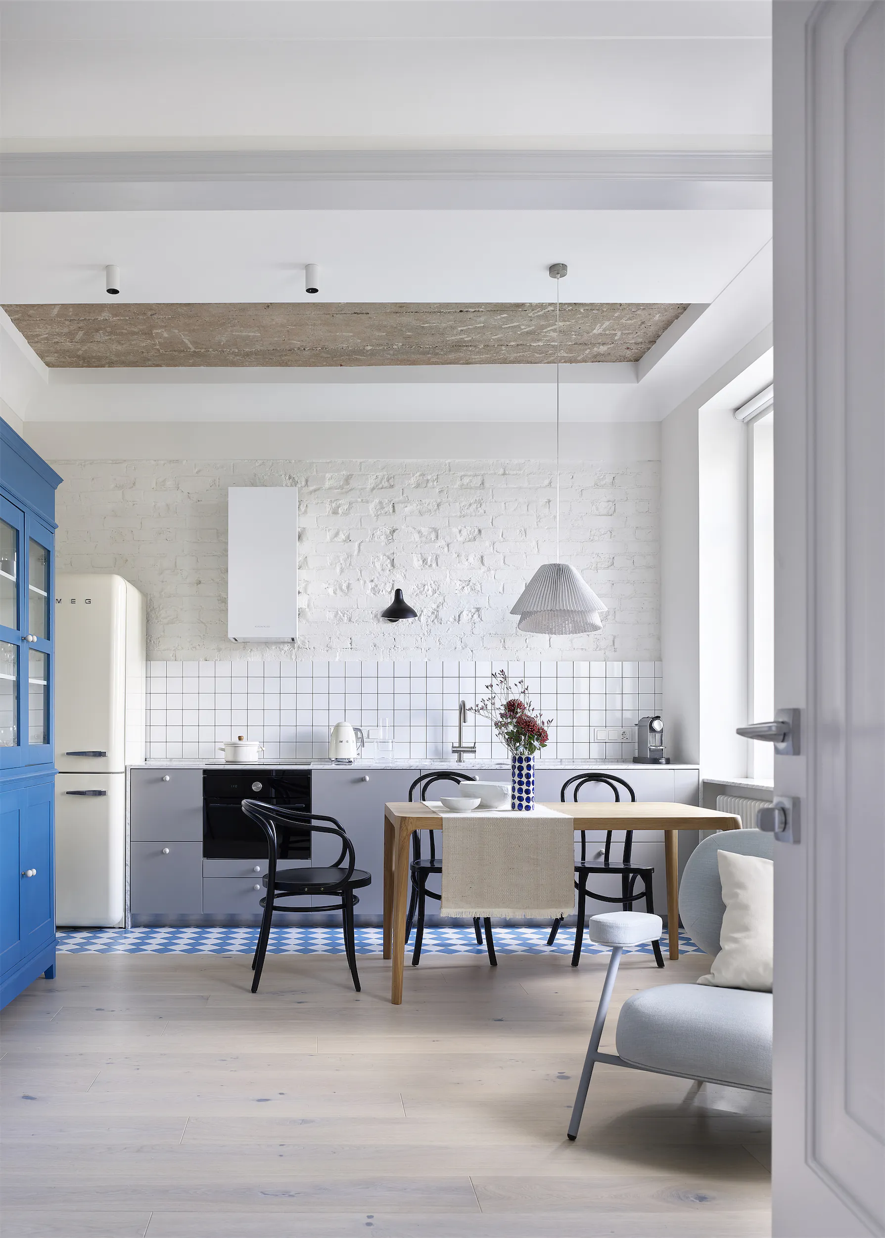 Минималистичная кухня с белыми стенами, деревянным столом, чёрными стульями, синим шкафом и белым холодильником.