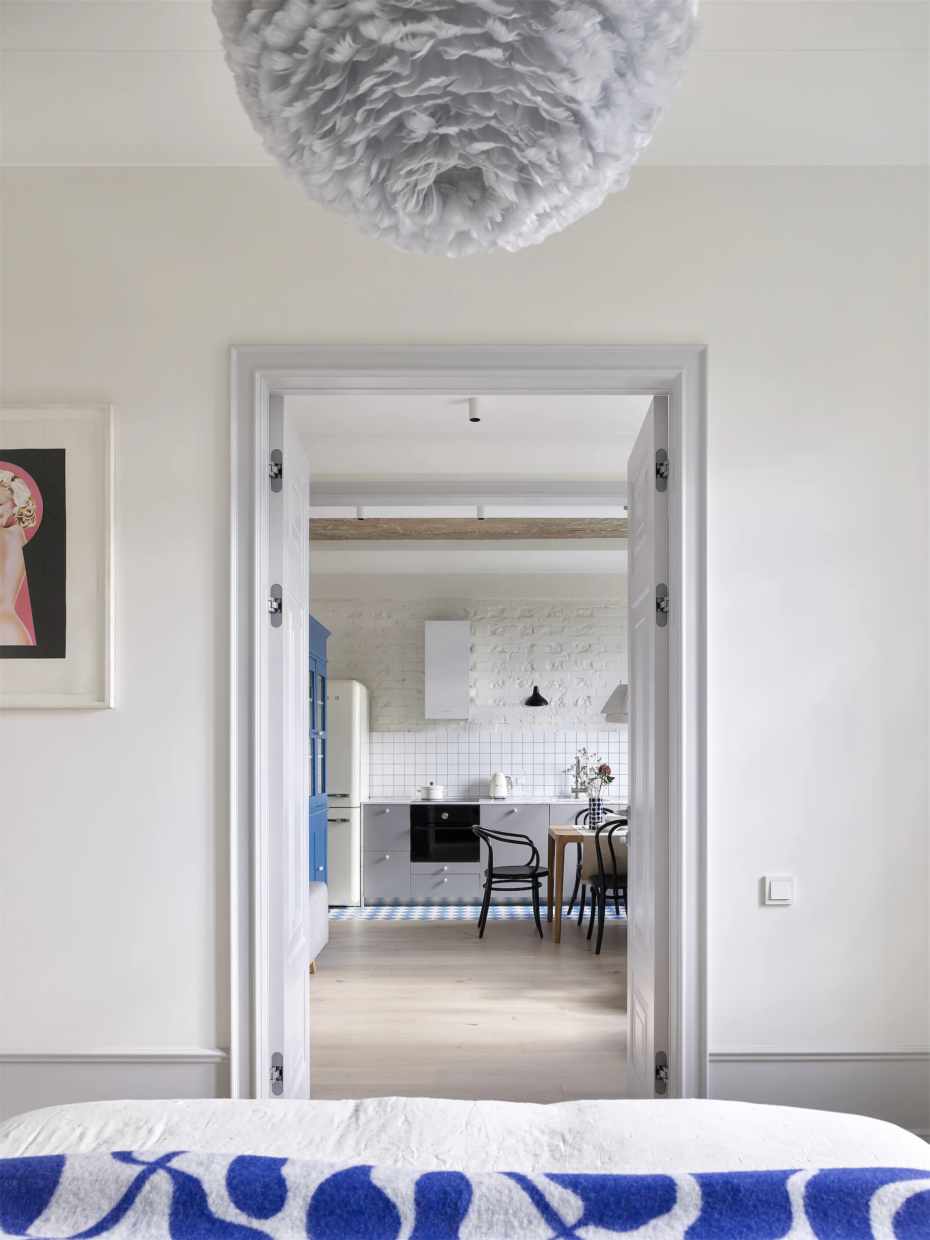 Вид из спальни через дверной проём на кухонно-столовую зону. Синее узорчатое покрывало на фоне белых стен.