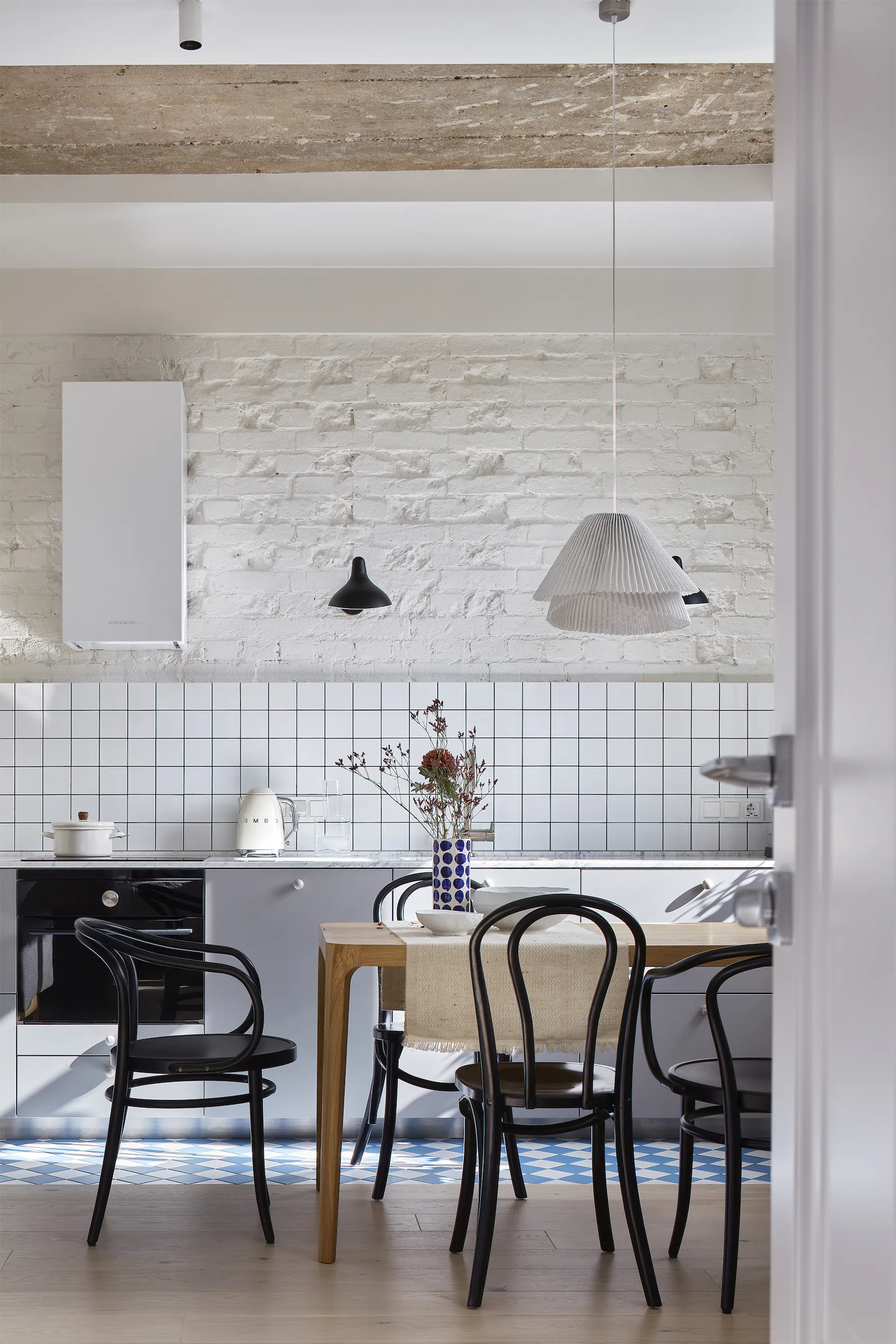 Минималистичная столовая с деревянным столом, чёрными стульями, белой кирпичной стеной и подвесным светильником.