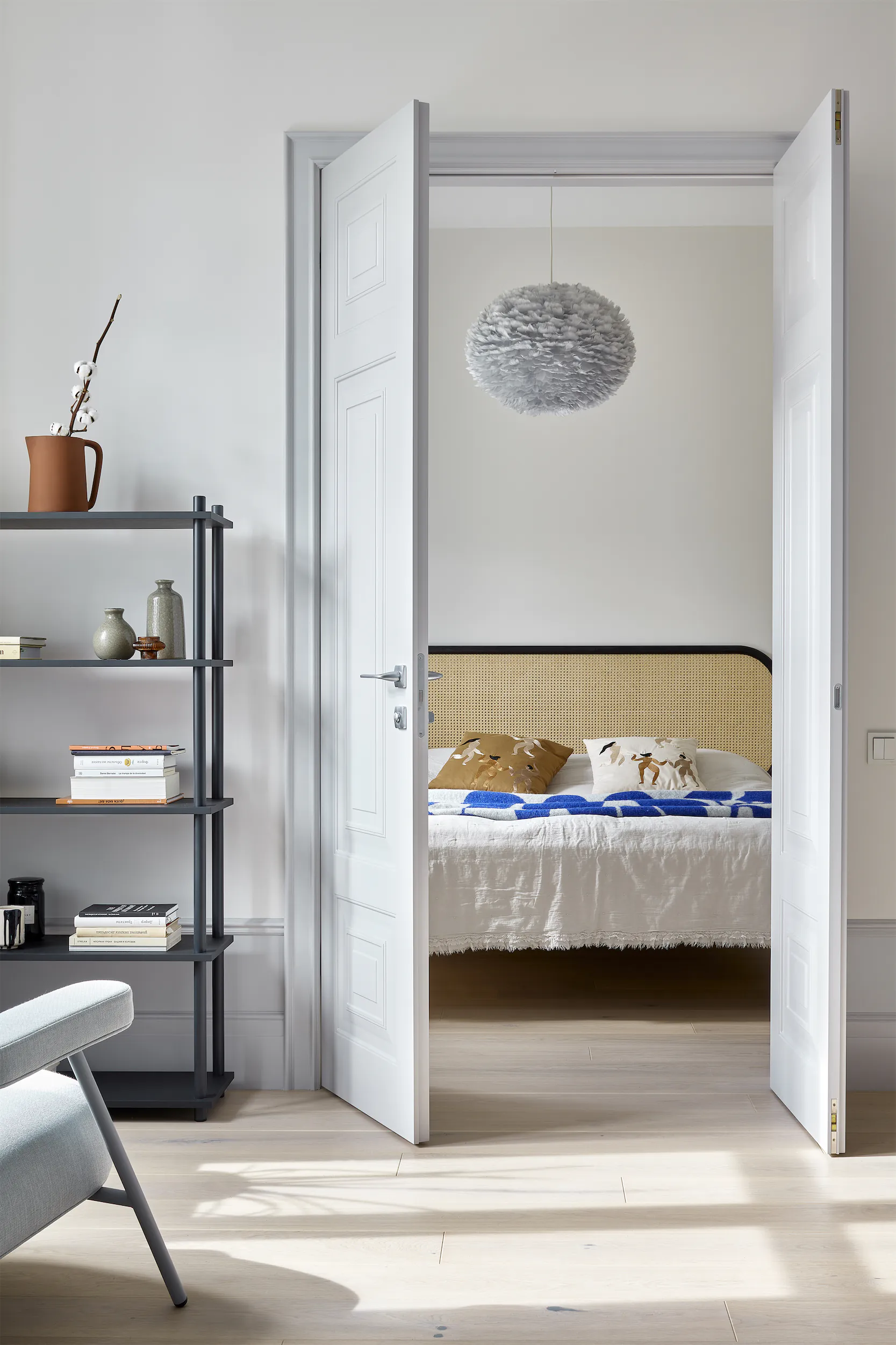 Открытая белая дверь открывает вид на спальню с кроватью, книжной полкой и мебелью.