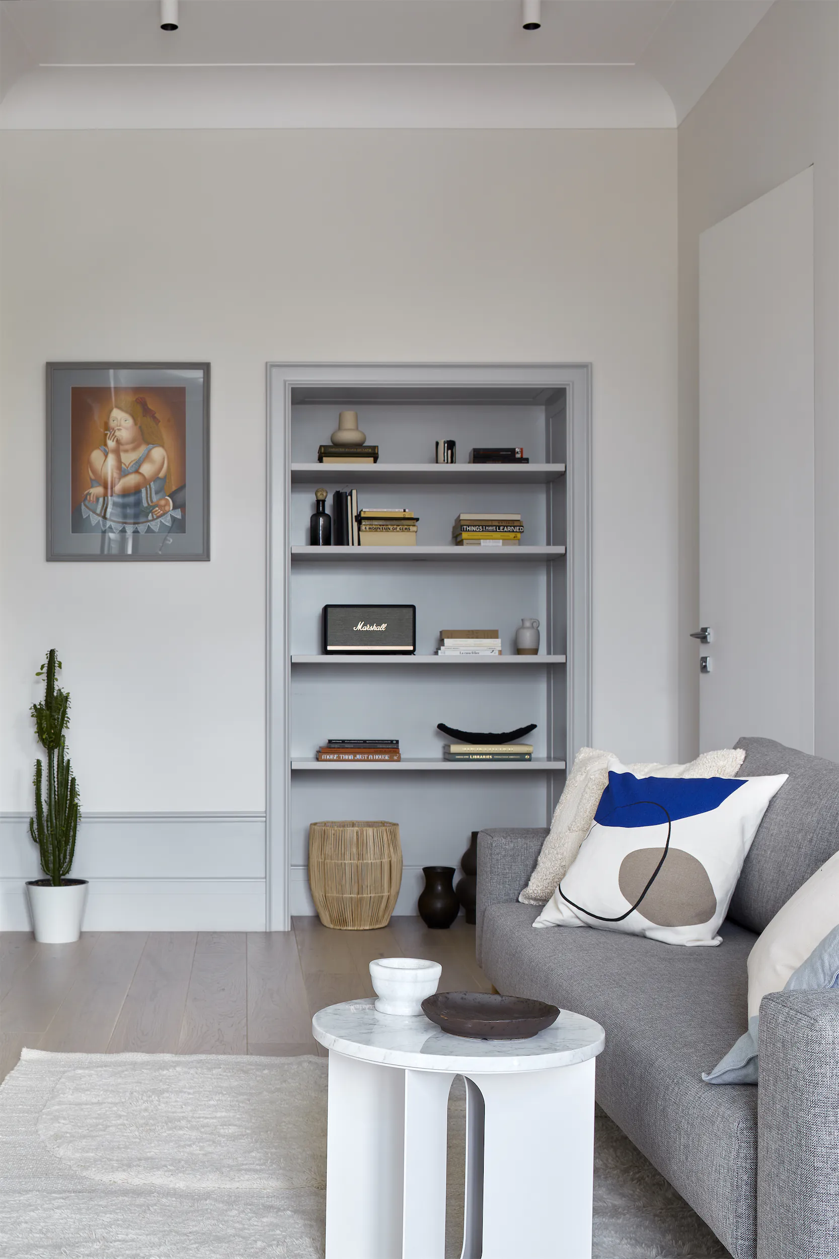 Минималистичная гостиная с серым диваном, белым столиком и книжной полкой у белой стены.