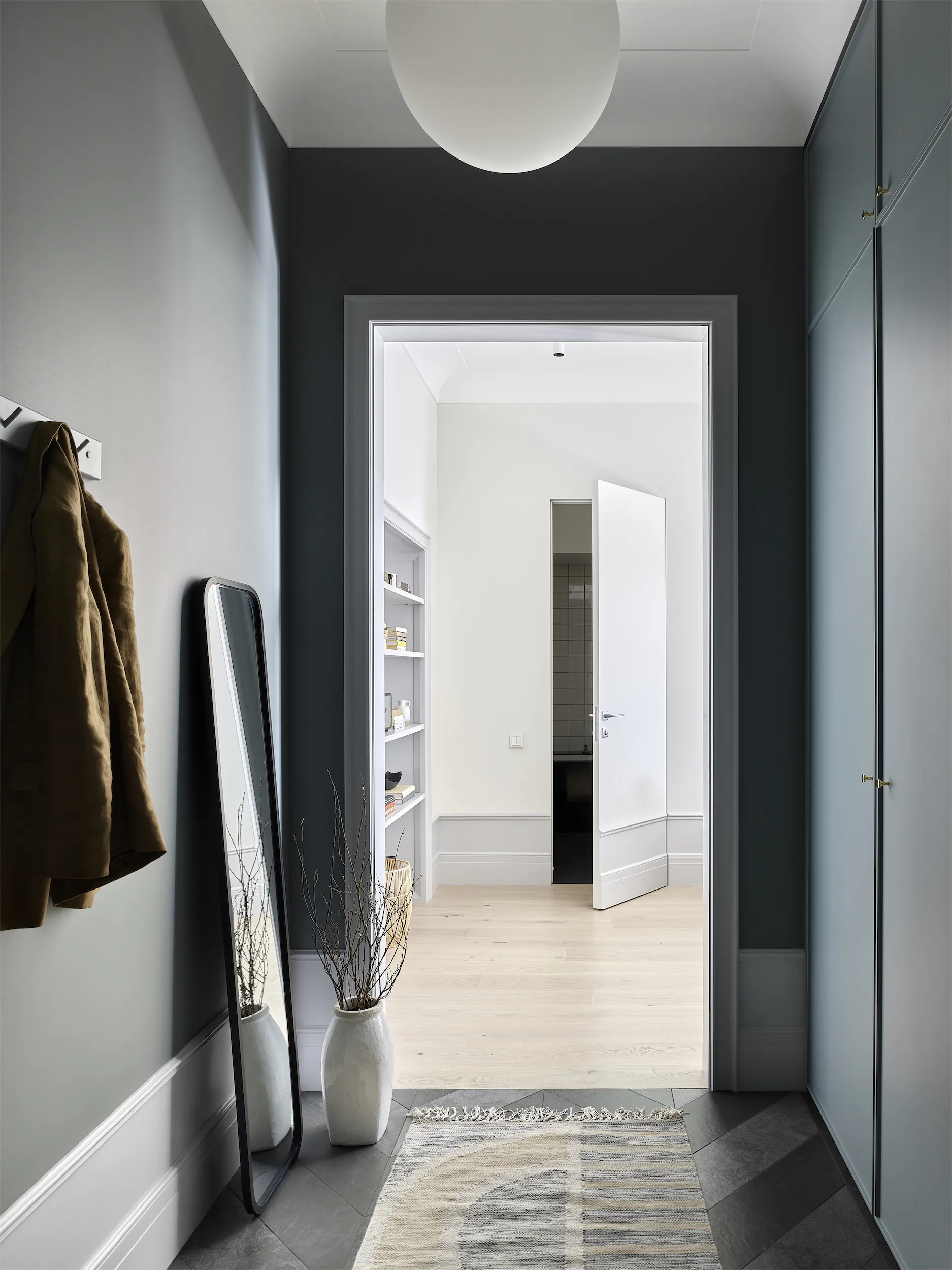 Коридор с серыми стенами, ведущий через белую дверь в комнату с книжной полкой и вазой на полу.
