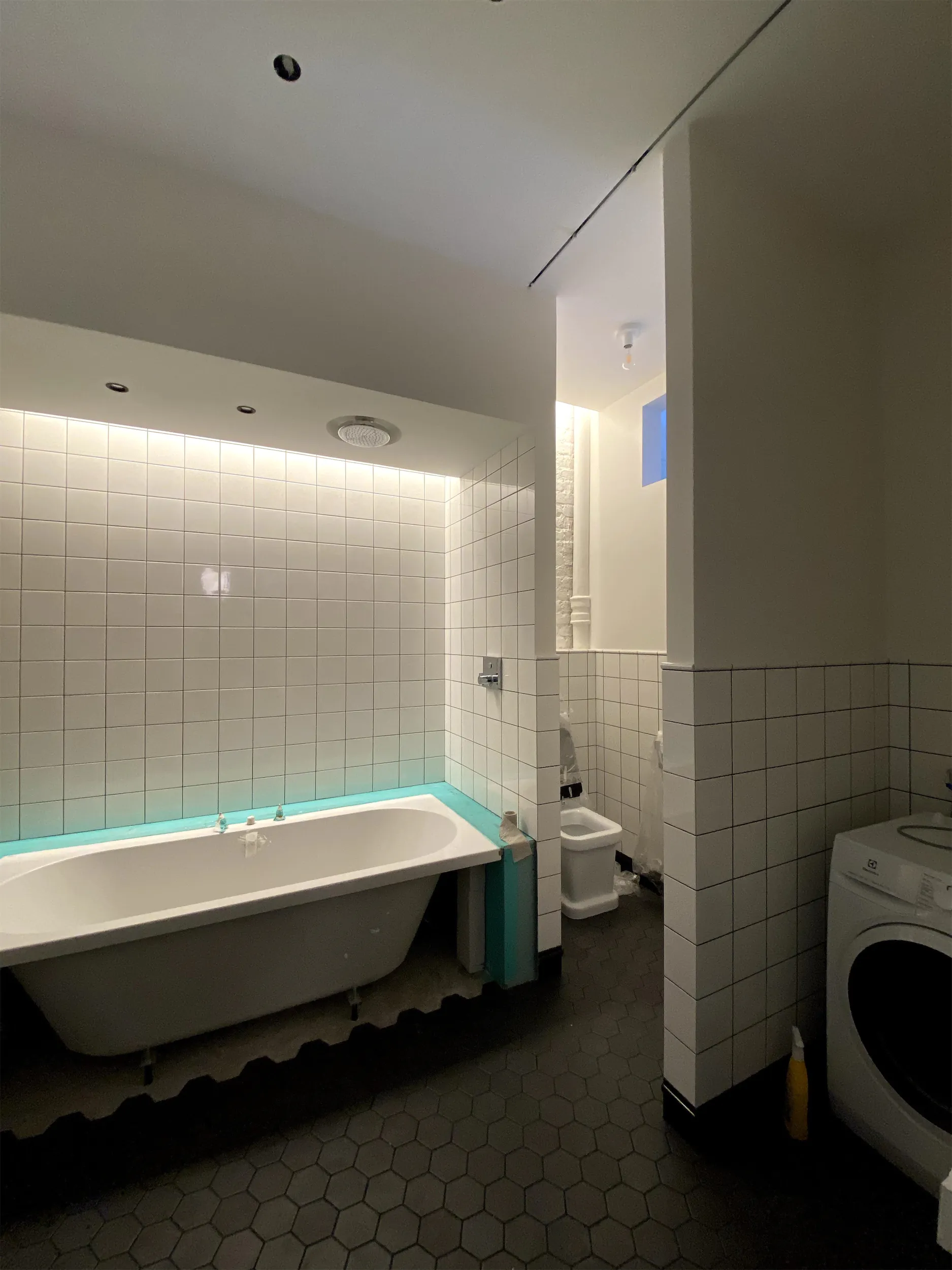 Ванная с ванной, унитазом и стиральной машиной. Стены из белой плитки, пол из тёмно-серой шестигранной плитки.