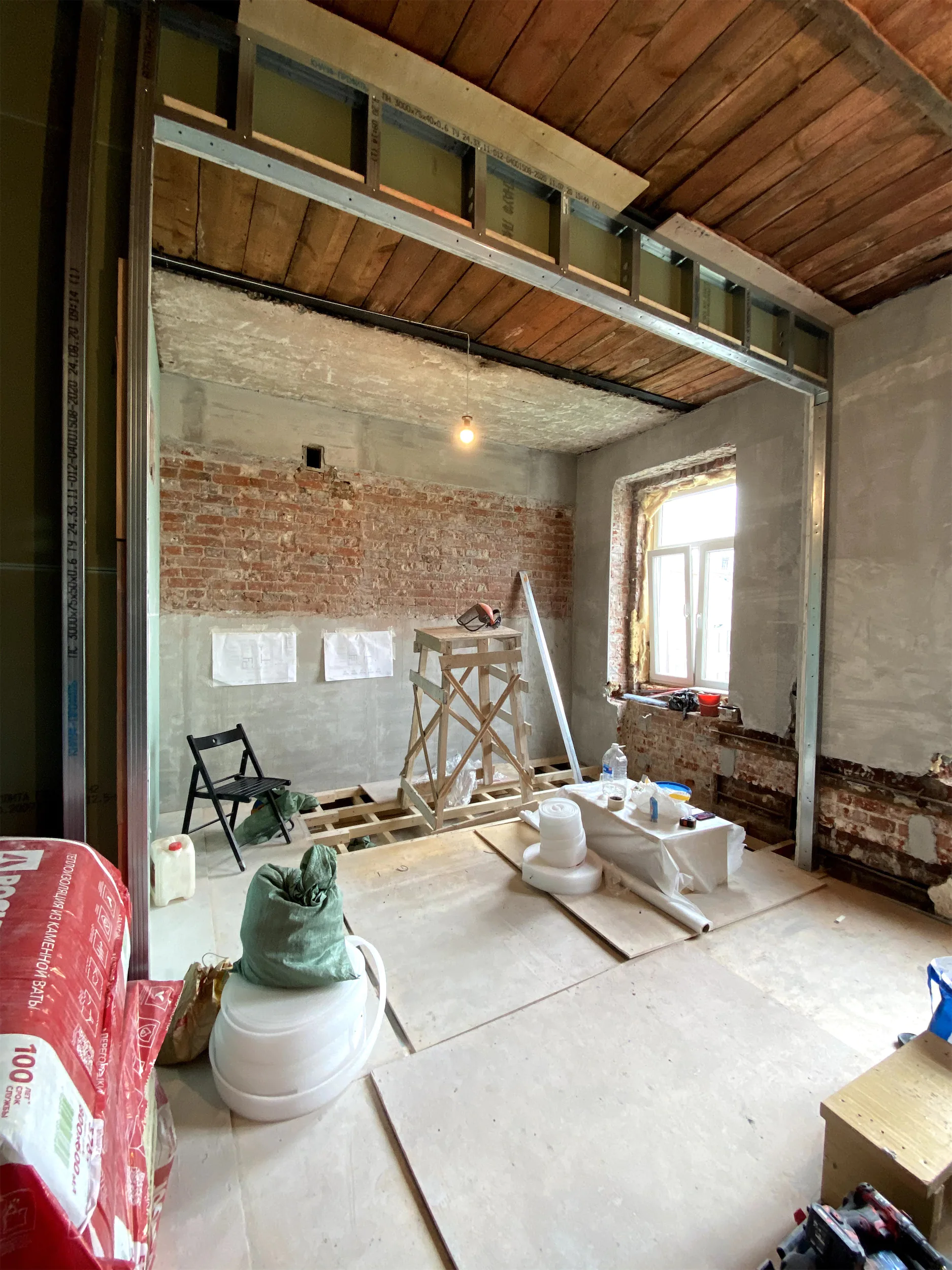 Комната в процессе ремонта с окном, открытой кирпичной кладкой и деревянным потолком.