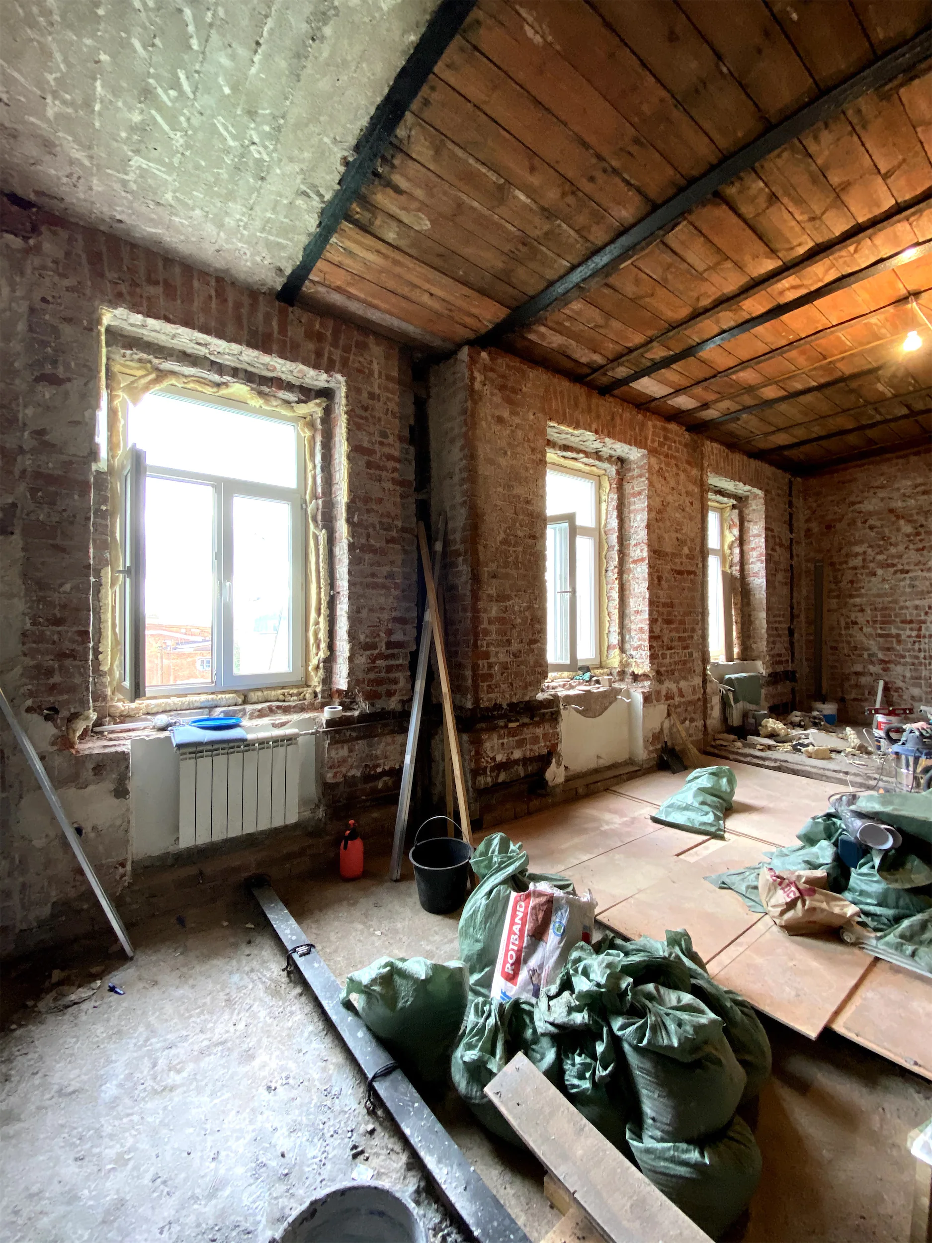Комната с открытой кирпичной кладкой и деревянным потолком. На полу разложена зелёная ткань.