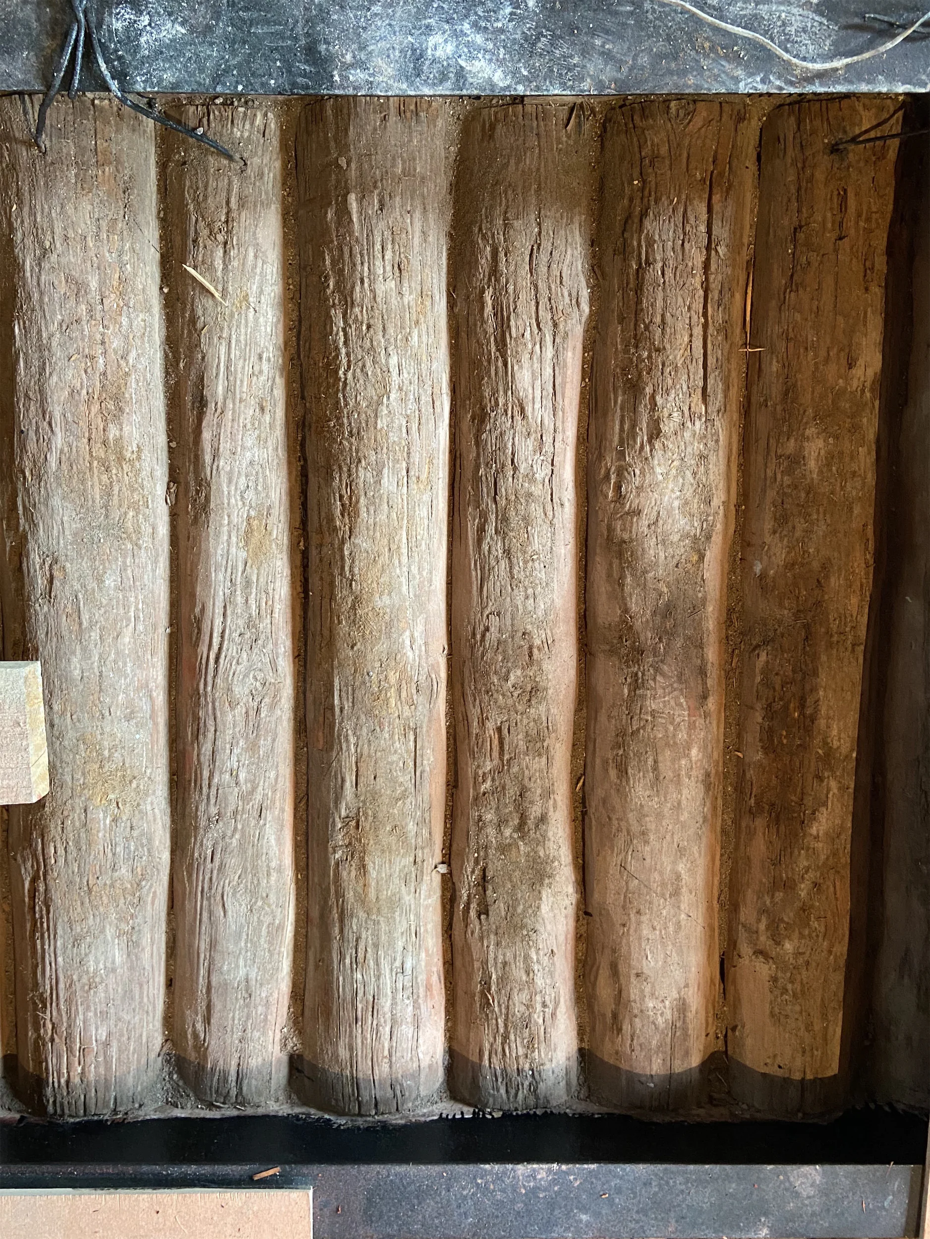 Стена из пяти вертикальных деревянных брёвен на тёмно-сером бетонном основании.