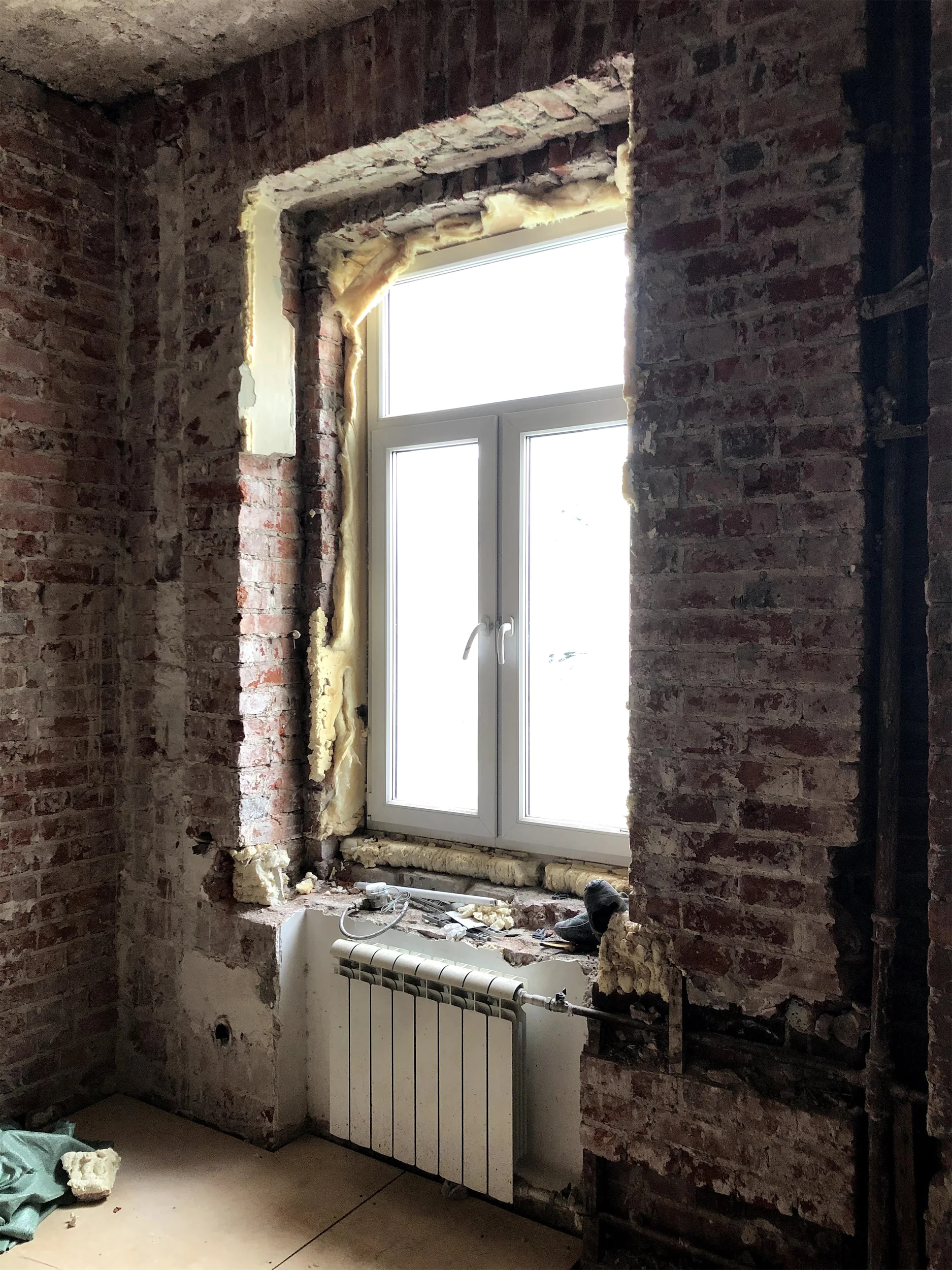 Комната с открытой кирпичной кладкой, радиатором и окном с белой рамой.