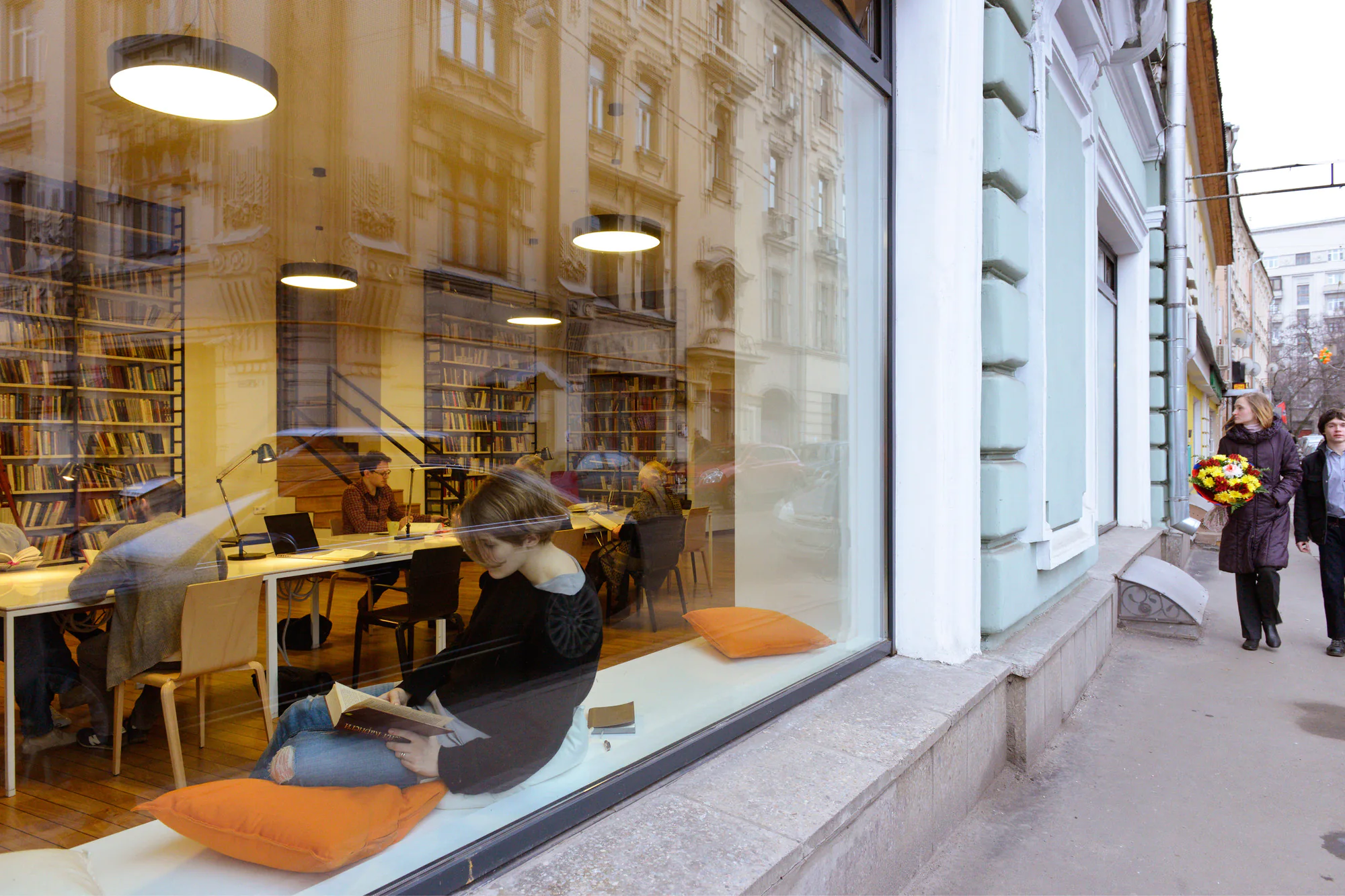 Женщина сидит на подоконнике с книгой среди стола и книжных полок.