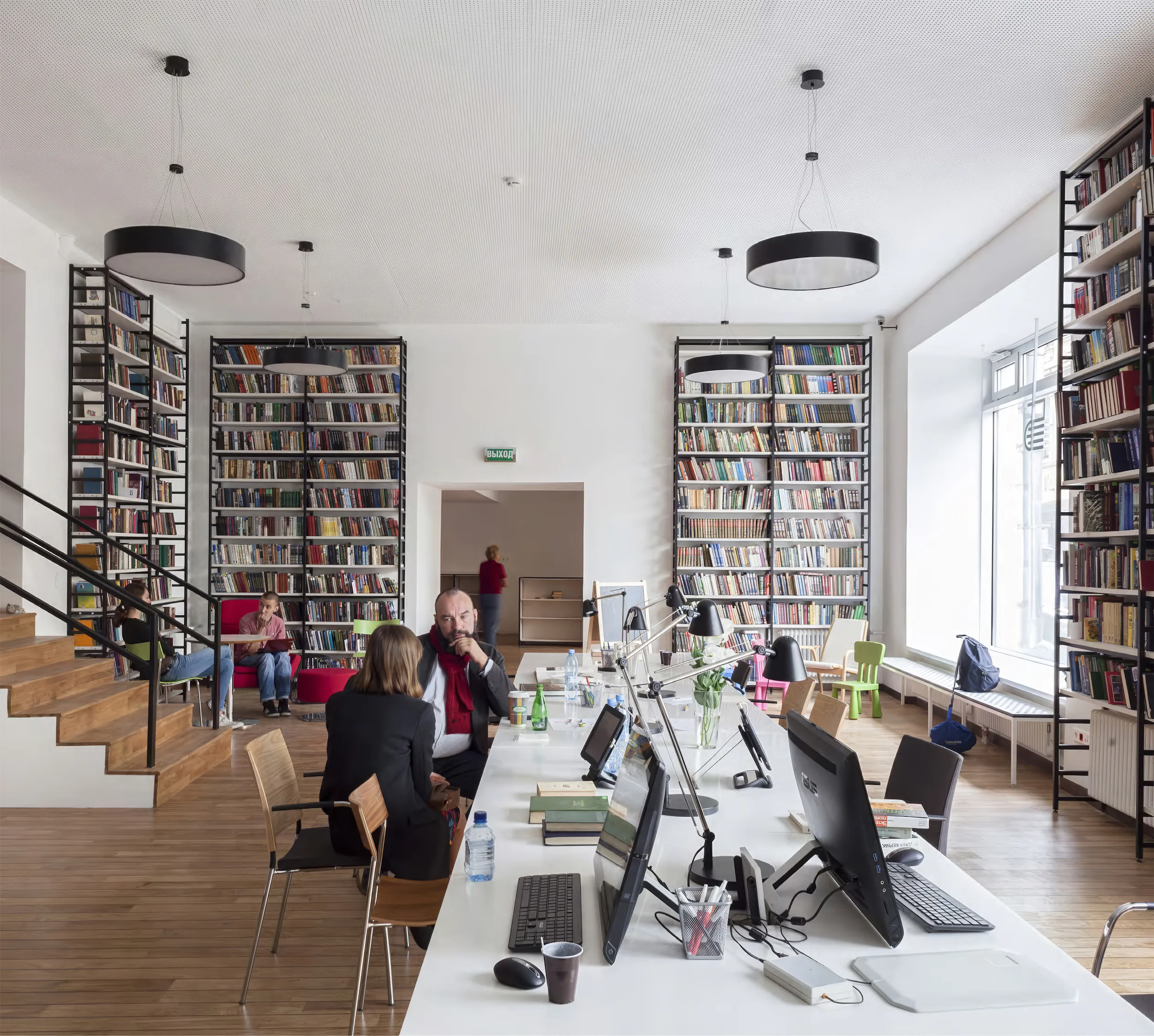 Офис с белым столом, книжными полками и лестницей. За столом сидят двое.