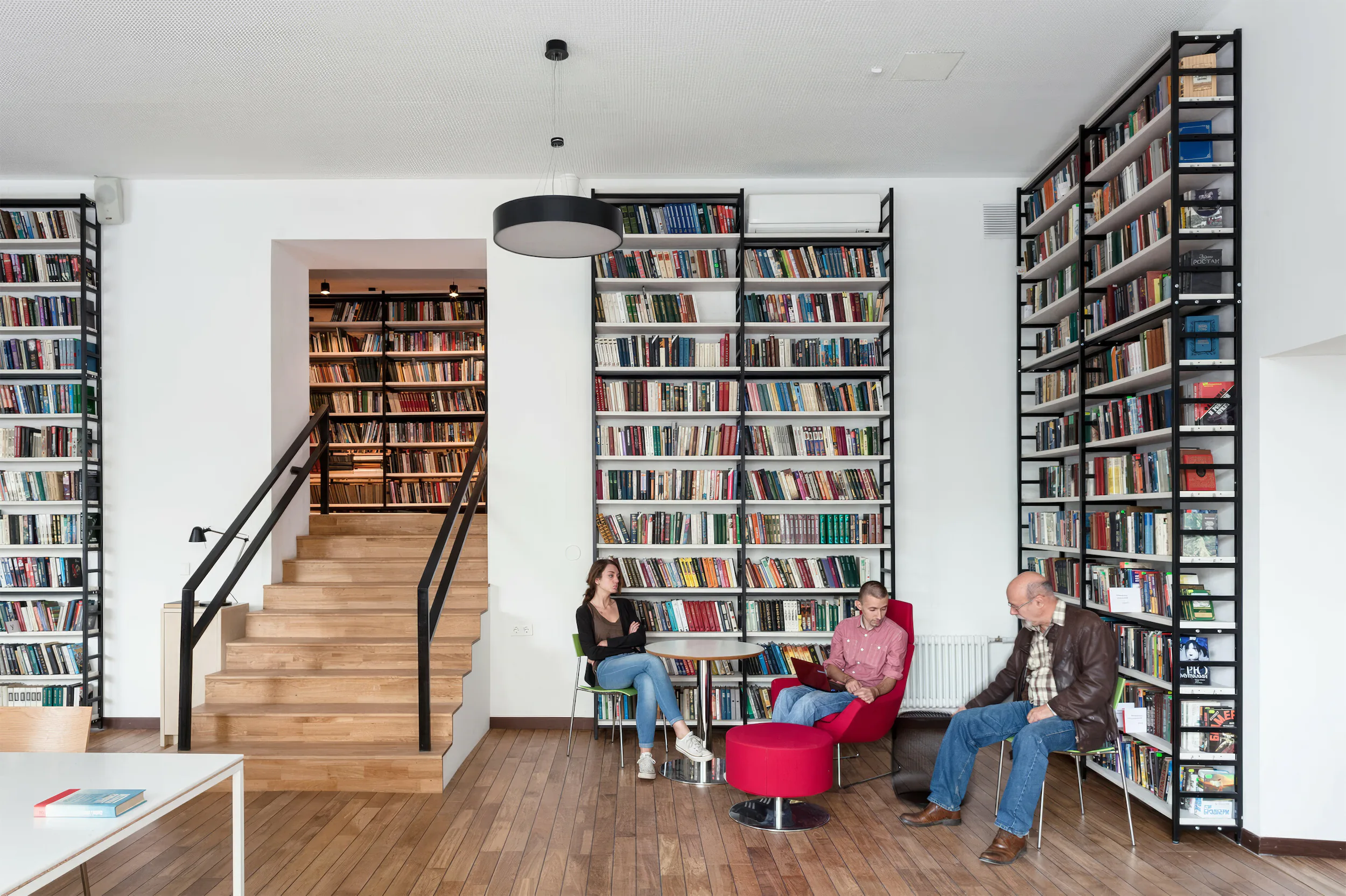 Современное пространство с лестницей и книжными полками. Двое сидят в красном и нейтральном кресле.