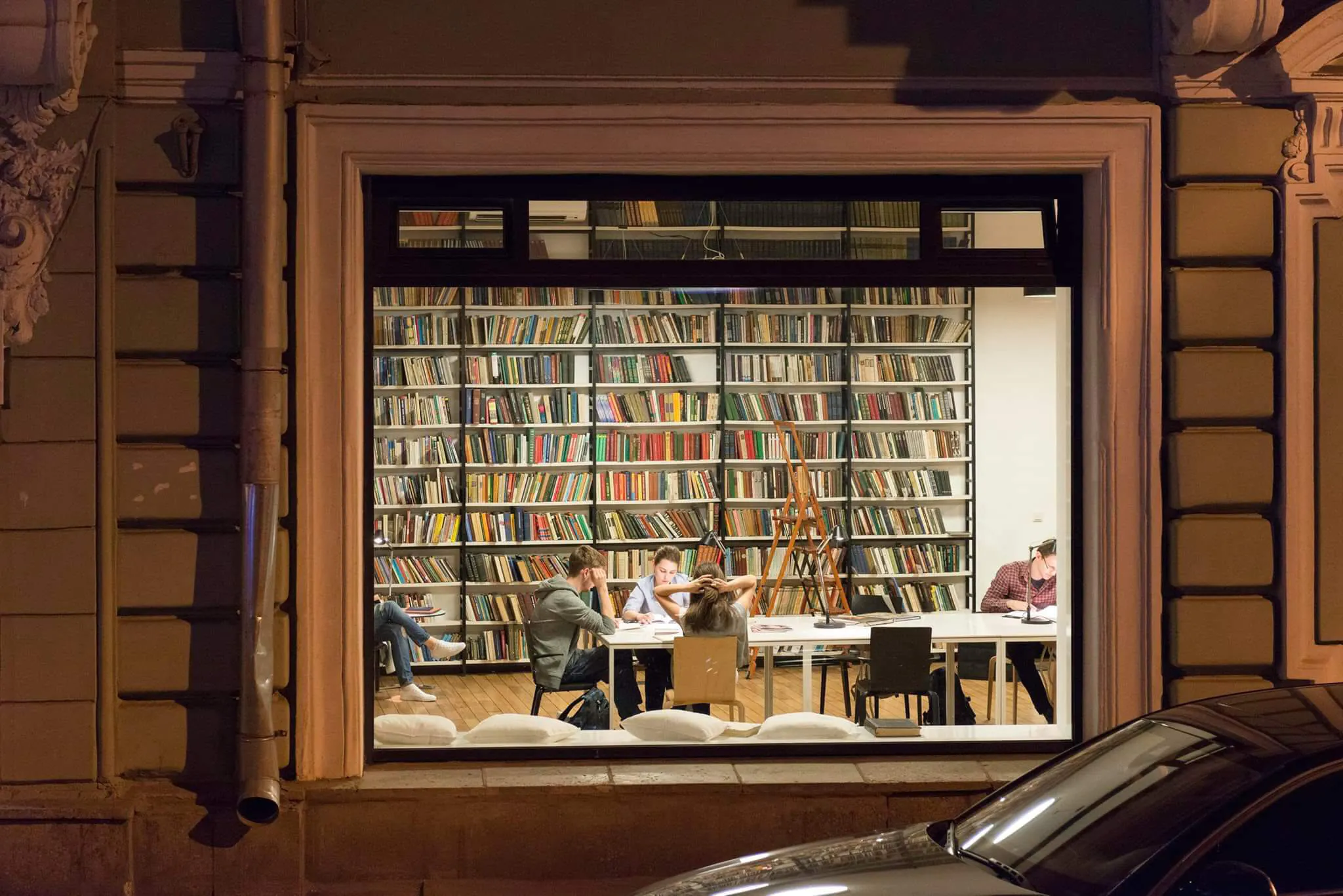 Окно с белой рамой, в котором отражаются книжная полка и сидящие люди.