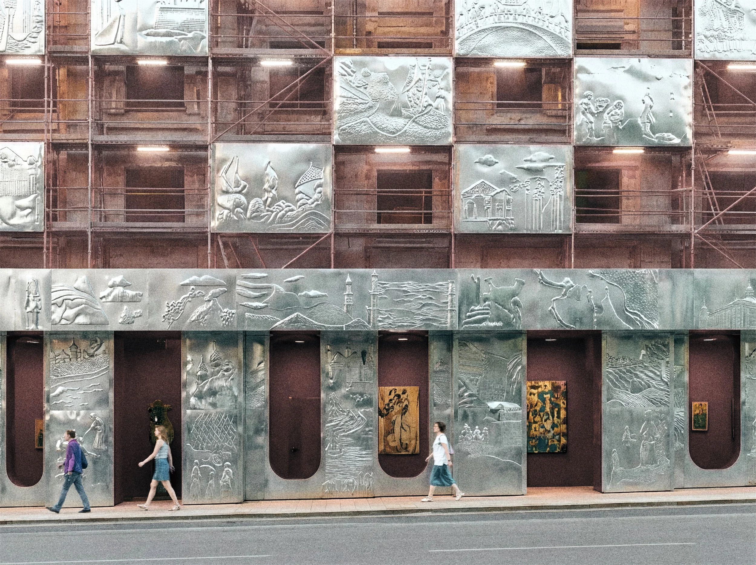A building facade with a metal exterior and intricate designs, with two people walking in front.