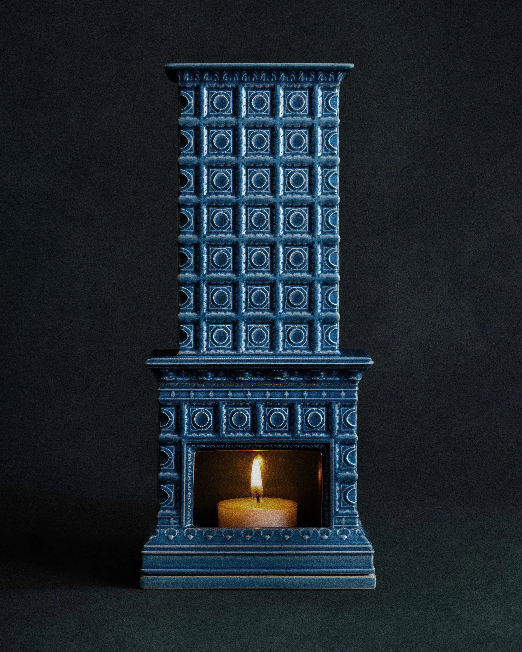 Подсвечник в форме синего камина с зажжённой свечой на тёмном фоне.