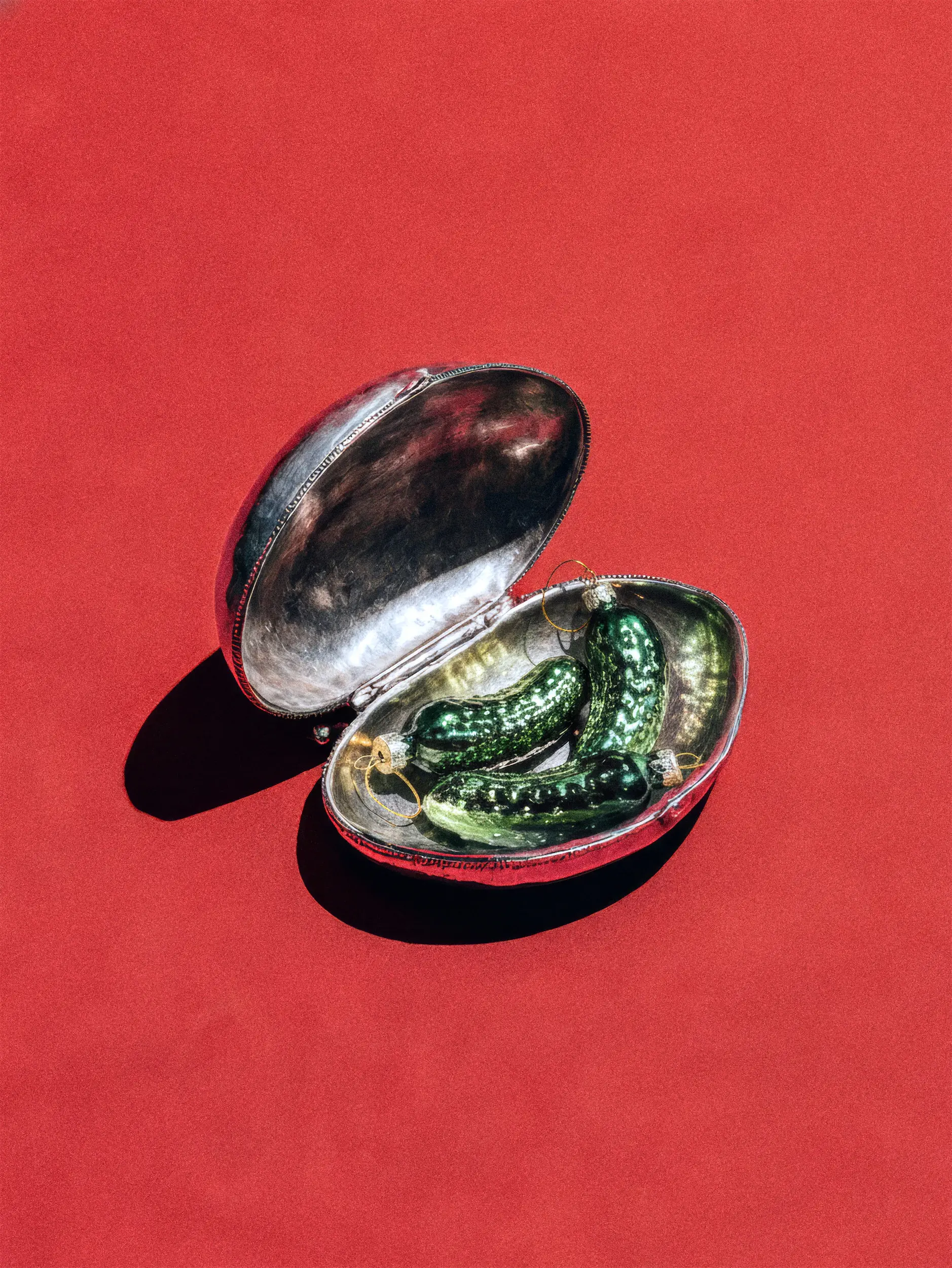 Серебряный овальный контейнер с откинутой крышкой. Внутри зелёный предмет, фон красный.