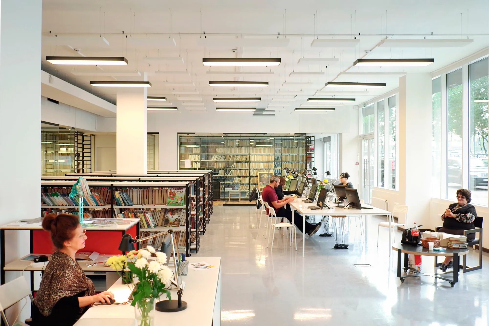 Современный офис с белым полом, большим окном и двумя людьми за столами. На заднем плане книжная полка.