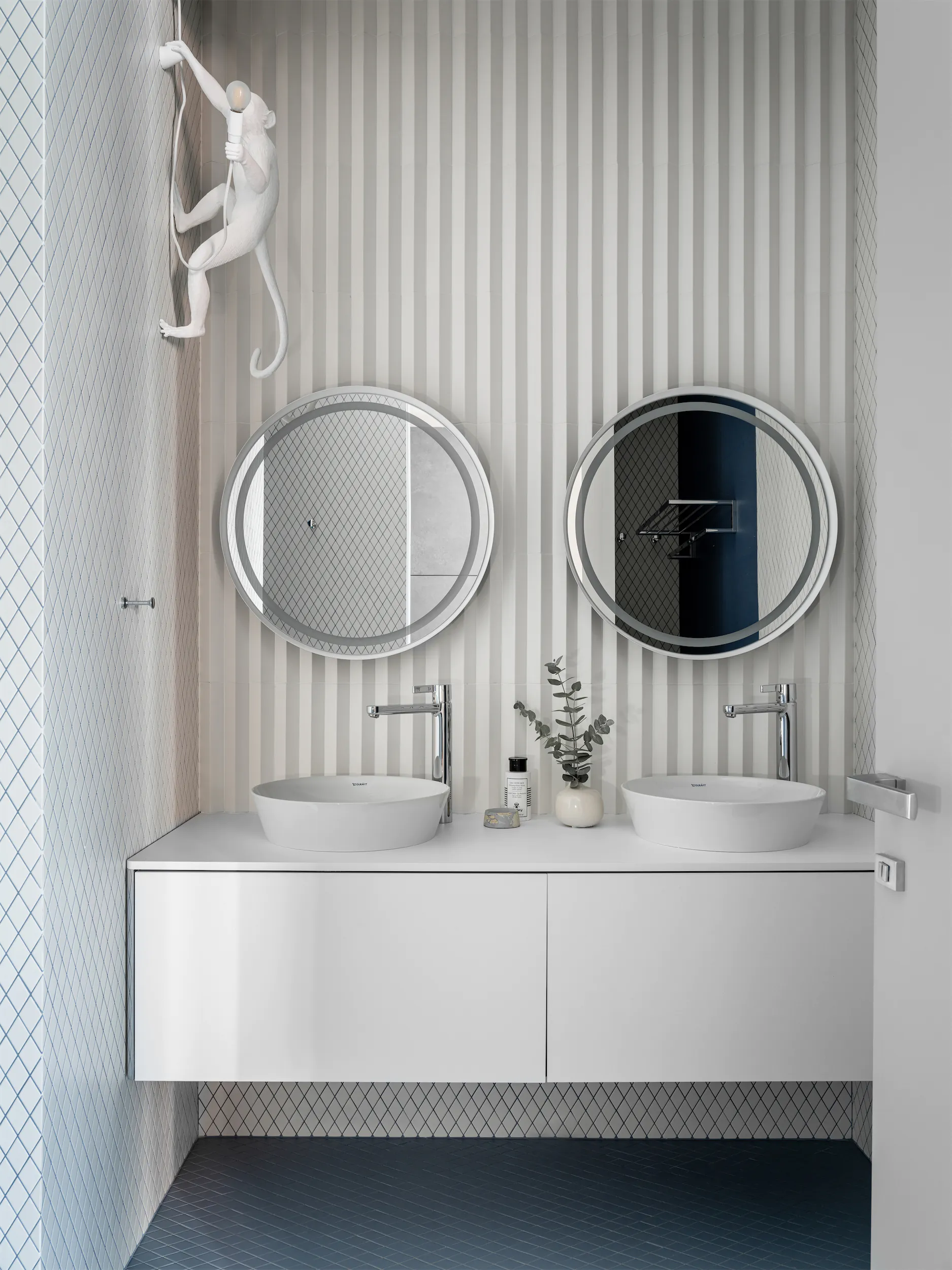 Современная ванная с двумя белыми раковинами, круглым зеркалом и скульптурой обезьяны на фактурной стене.