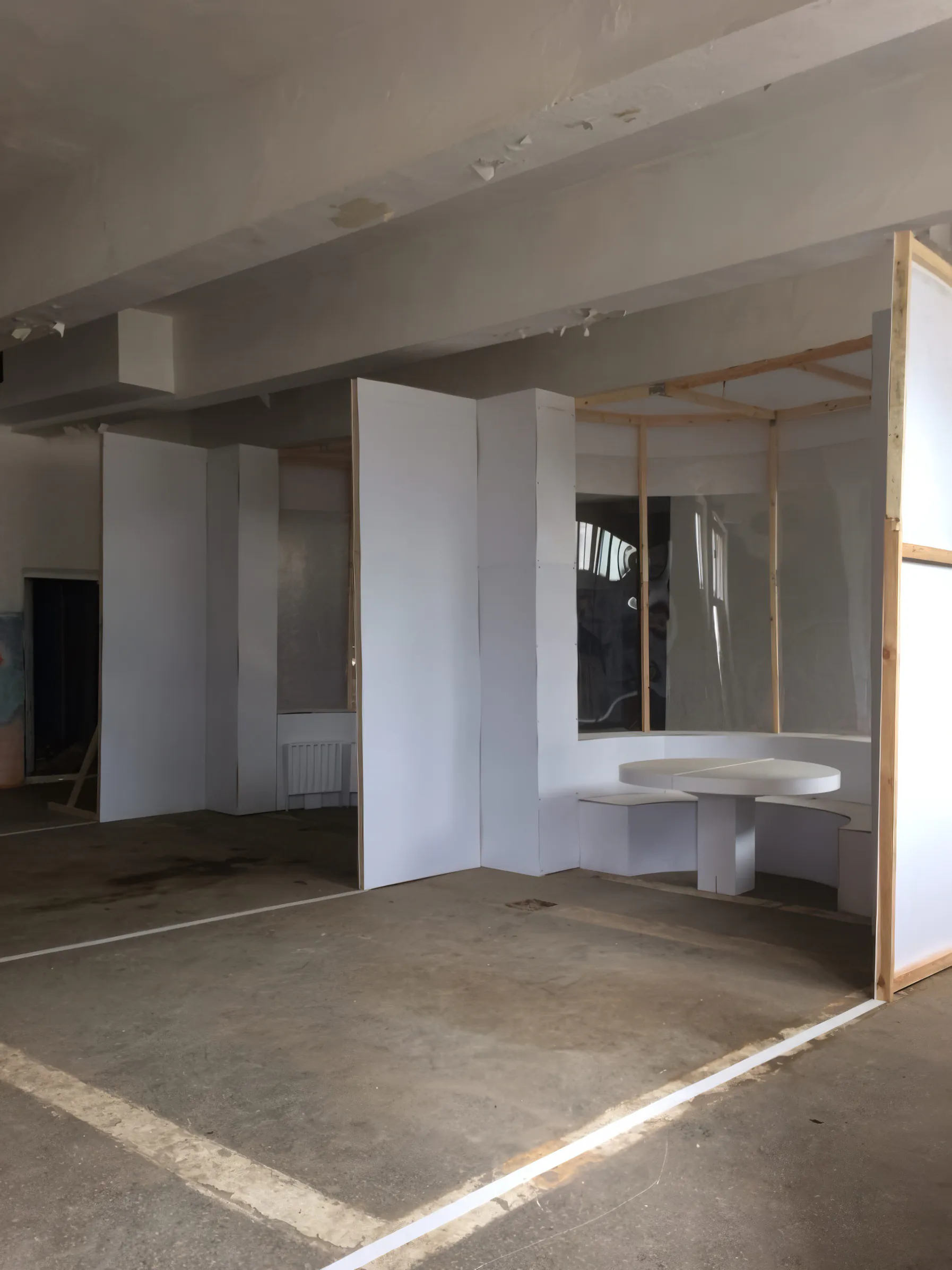 Комната с белыми стенами, бетонным полом и дверным проёмом с деревянным обрамлением.